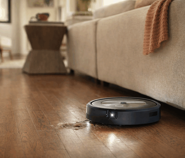 Robot vacuum cleans hardwood floor beside sofa in sunlit living room, collecting dust under furniture with modern home setting interior.