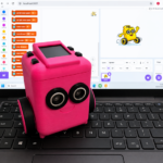 Small pink robot sits on a laptop keyboard in front of a coding screen showing a Scratch style programming interface