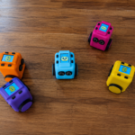 Five colourful Tinkimo robots sit on a wooden floor, each displaying expressive animated faces while spaced apart in a playful formation.