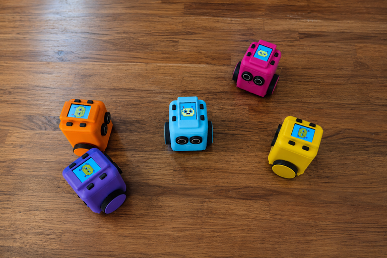 Five colourful Tinkimo robots sit on a wooden floor, each displaying expressive animated faces while spaced apart in a playful formation.